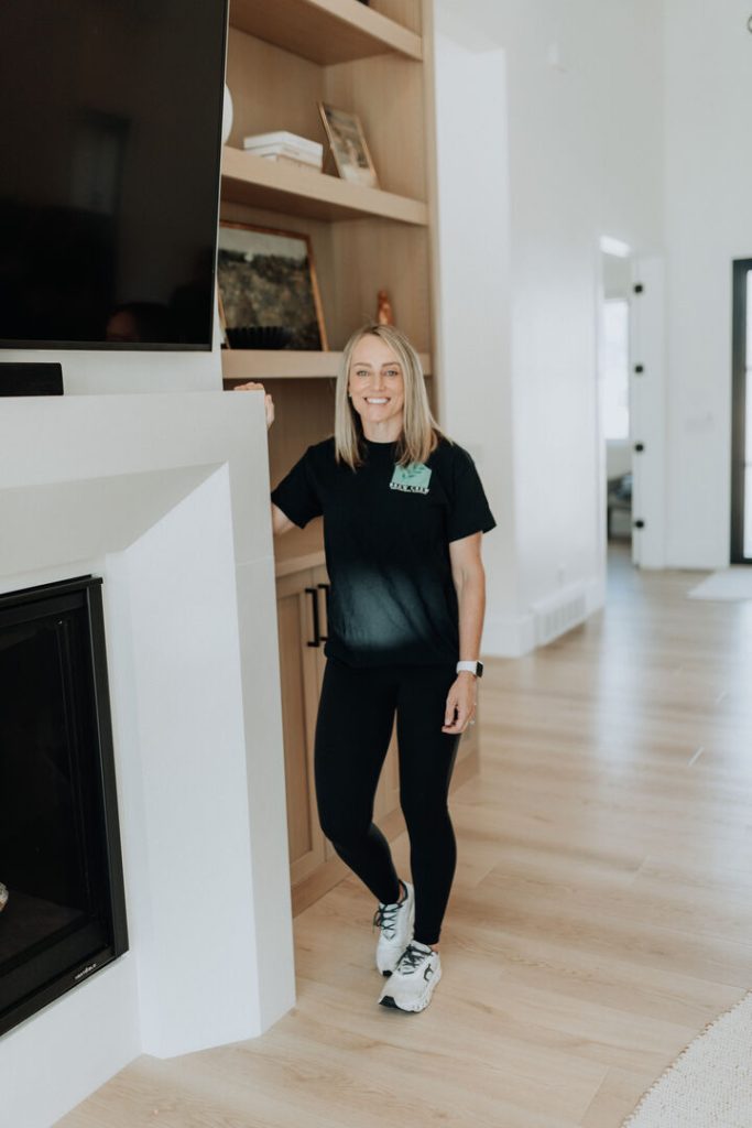 Smiling owner stands beside a modern fireplace and wooden shelves in a bright, welcoming home.