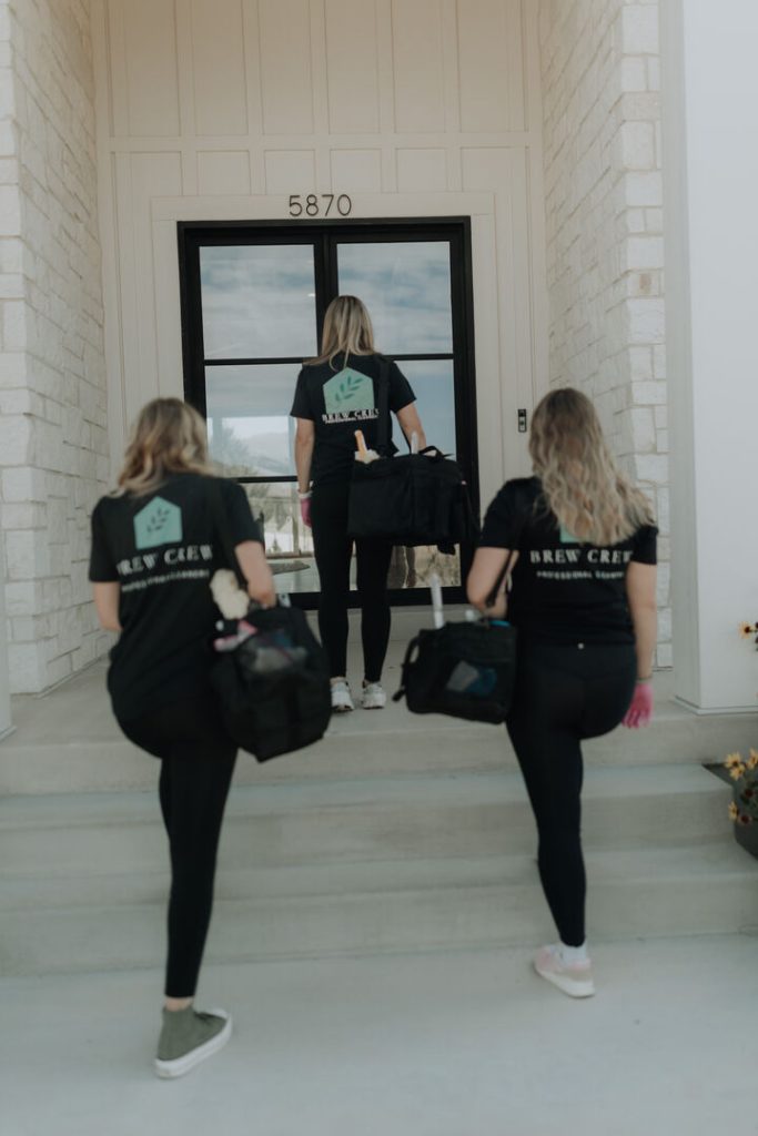 Three women in matching black “Brew Crew” shirts carry coffee gear up white stone steps toward a glass entry marked 5870, creating a focused, welcoming team atmosphere.