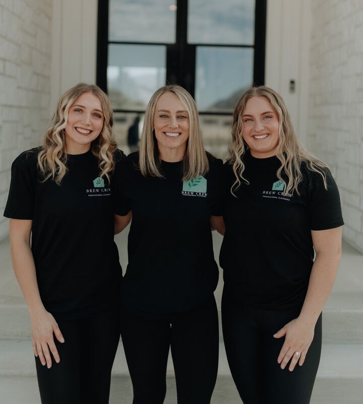 Three smiling women in matching black shirts stand arm-in-arm on steps inside a bright, white-brick entryway, projecting warmth and team spirit.