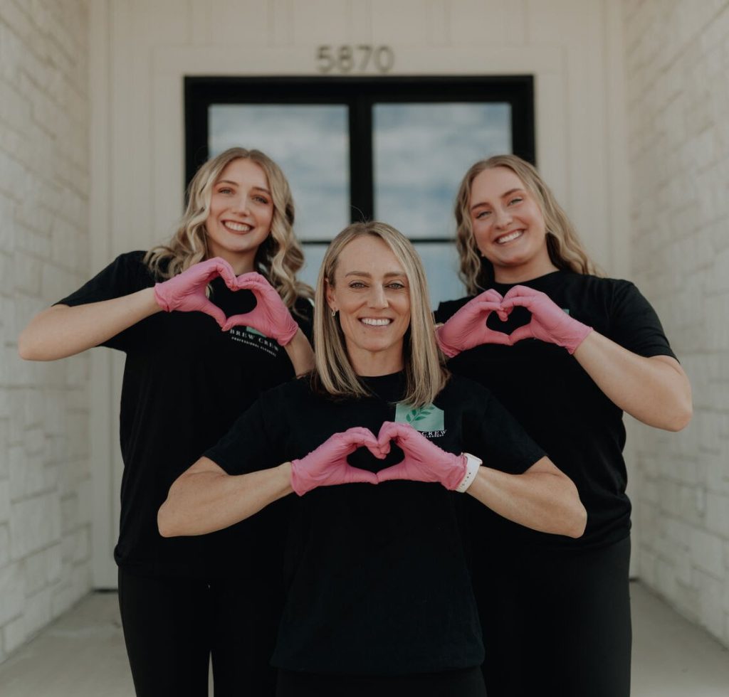 Three smiling women in black outfits and pink gloves form heart shapes with their hands outside a stone building entrance, conveying warmth and teamwork.